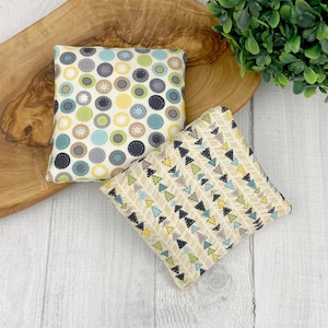 May include: Two small, square fabric items with patterned designs. One features circles in blue, green, yellow, and gray with stitched details on a cream background. The other has a repeating triangle pattern in similar colors on a tan background.