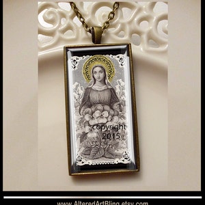May include: A vintage-style pendant necklace featuring a black and white image of a woman holding flowers. The image is framed in a gold-toned metal frame and set against a white lace background. The pendant is on a gold chain.