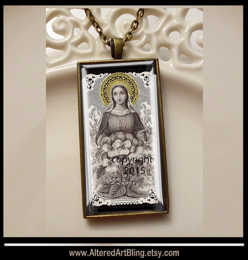 May include: A vintage-style pendant necklace featuring a black and white image of a woman holding flowers. The image is framed in a gold-toned metal frame and set against a white lace background. The pendant is on a gold chain.