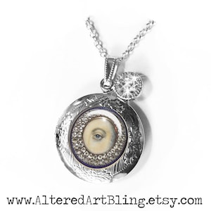 May include: Silver locket necklace with a painted eye design and a small crystal charm. The locket is open and the eye is visible.