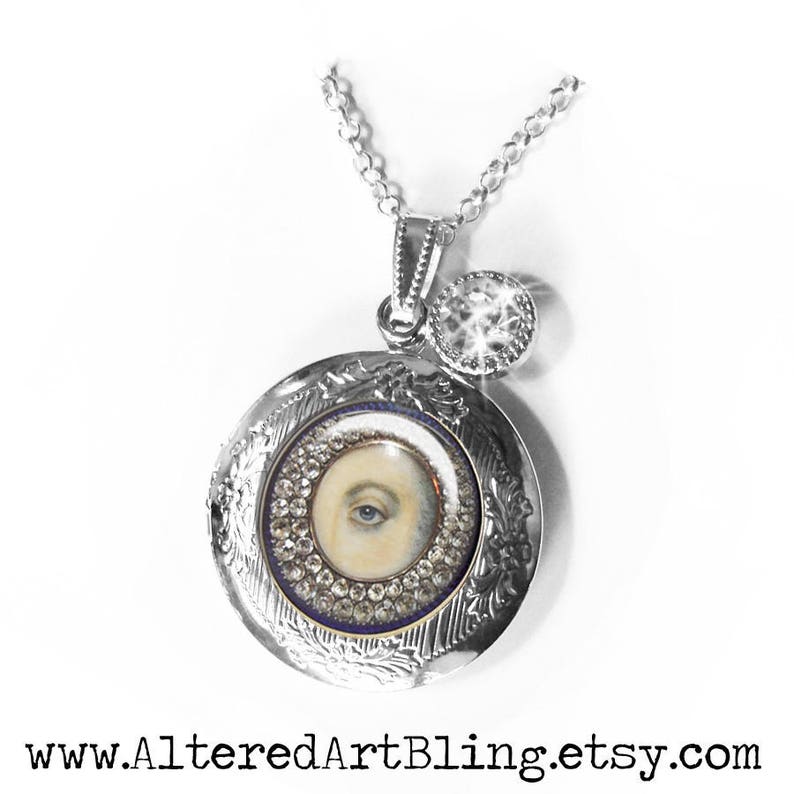 May include: Silver locket necklace with a painted eye design and a small crystal charm. The locket is open and the eye is visible.