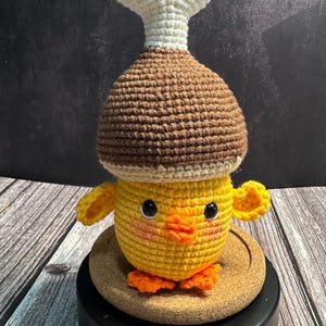 May include: A crocheted chick toy, featuring a brown hat and a white bone-shaped top. The chick is yellow with orange feet and wings. The toy is displayed on a cork base.