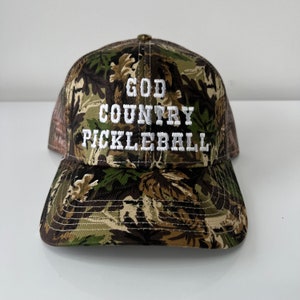 May include: A camouflage trucker hat with the text "GOD COUNTRY PICKLEBALL" embroidered in white.