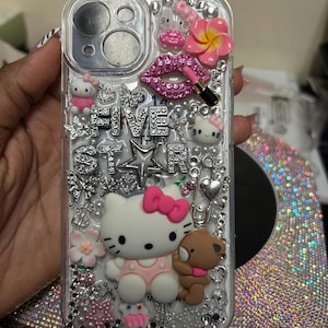 Puede incluir: Funda transparente para teléfono decorada con Hello Kitty, un oso pardo, labios rosas y un lápiz labial. La funda presenta las palabras "FIVE STAR" en plata, rodeadas de pedrería, perlas y otros adornos. También se ven una flor rosa y una magdalena.