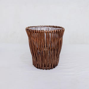 Może przedstawiać: Brązowy, pleciony kosz z wikliny z przezroczystą plastikową wkładką. Kosz jest cylindryczny z lekko rozszerzonym górnym brzegiem. Splot jest pionowy, tworząc teksturowaną powierzchnię. Kosz nadaje się do roślin lub przechowywania.