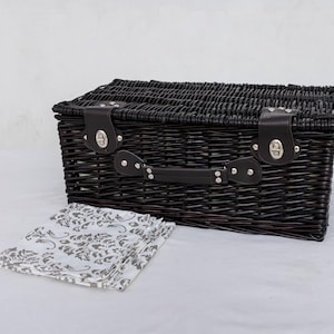 May include: A black wicker picnic basket with a matching handle and leather straps. The basket is closed and has silver-colored clasps. A folded white and gray patterned cloth is in front of the basket.
