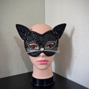 May include: A black cat-eye mask with sequin and bead embellishments. The mask features cat ears, a rhinestone nose, and silver whiskers. The mask is displayed on a mannequin head.