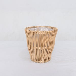 May include: A light brown wicker basket, cylindrical in shape, with a woven design. The basket has a clear plastic liner inside. The basket is made of natural materials and is suitable for home decoration.