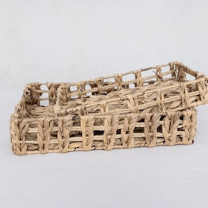 Puede incluir: Dos cestas rectangulares tejidas apiladas una encima de la otra. Las cestas están hechas de material tejido de color natural con un diseño de tejido rectangular abierto. Las cestas están vacías y se encuentran sobre una superficie blanca.
