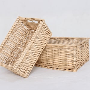 May include: Two rectangular woven wicker baskets. One basket is upright, and the other is leaning against it. The baskets are a light tan color and have handles. The background is a plain white.