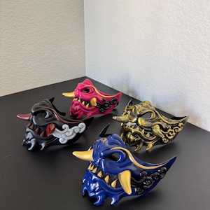 May include: Four colorful Oni masks are displayed on a dark surface. The masks are in black, pink, gold, and blue, each with unique details like horns, teeth, and cloud-like accents. The masks are likely for cosplay or decorative purposes.