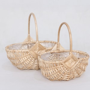 May include: Two woven wicker baskets with arched handles. The baskets are oval-shaped and lined with clear plastic. The wicker has a light brown color. The baskets are empty and ready to be filled.