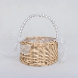 May include: A small, round woven basket with a handle adorned with white faux pearls. The basket is decorated with white sheer ribbons tied in bows. The basket is made of light brown wicker and is lined with clear plastic.