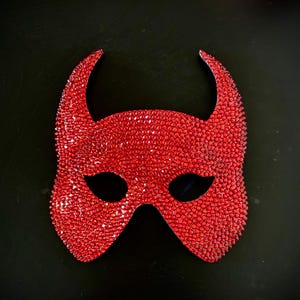 May include: A red devil mask with curved horns and eye openings. The mask is covered in small, sparkling red rhinestones. The mask is set against a black background.