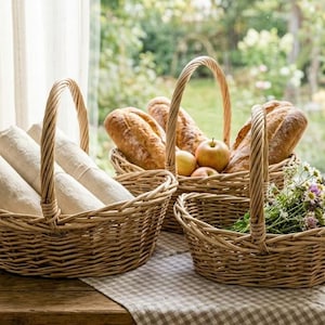 May include: Three woven wicker baskets on a wooden table. One basket holds rolled white linens, another contains loaves of bread and apples, and the smallest has flowers. A mug sits nearby.