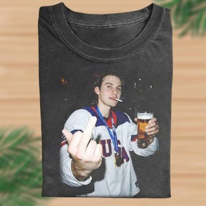 May include: Black t-shirt featuring a photograph of a person holding a beer and cigarette, wearing a white and blue jersey with a "USA" pendant. The person is giving the middle finger. The background is dark.