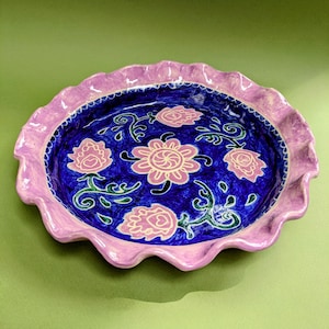 Puede incluir: Un plato de cerámica redondo con un borde ondulado de color morado claro. El centro presenta un esmalte azul intenso decorado con una flor rosa central rodeada por cuatro motivos florales rosas más pequeños y patrones de enredaderas verdes.