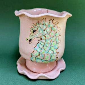 May include: A light pink ceramic planter with a ruffled rim and matching saucer. The exterior features a hand-painted seahorse illustration in shades of light green and teal, outlined in brown, set against the smooth, pale pink glazed surface.