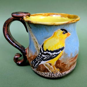 May include: A ceramic mug featuring a hand-painted yellow goldfinch with black and white wing markings against a light blue background. The mug has a dark brown, curved handle and a bright yellow interior glaze with a brown rim.
