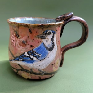 Puede incluir: Una taza de cerámica con esmalte moteado rosa y naranja que presenta un arrendajo azul pintado a mano posado en una rama. La taza tiene un asa marrón curva con un pequeño apoyo circular para el pulgar en la parte superior y forma redondeada.
