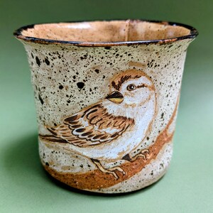 May include: A ceramic mug with a speckled cream-colored glaze and a dark rim. The exterior features a hand-painted illustration of a brown and white sparrow perched on a branch. The mug has a wide, flared opening and a cylindrical body shape.