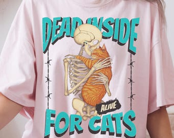 May include: A pink oversized t-shirt with a graphic print of a skeleton holding a cat. The text on the shirt reads "Dead Inside Alive For Cats".