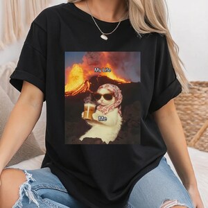 May include: Black t-shirt with a graphic of a cat wearing sunglasses and a headscarf, holding a beer. The text "My Life" is above the cat and "Me" is below the cat. The cat is sitting in front of a volcano.