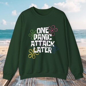 Könnte beinhalten: Ein dunkelgrüner Pullover mit dem Text "ONE PANIC ATTACK LATER" in weißen Buchstaben. Der Text ist von bunten, blumenförmigen Designs umgeben.