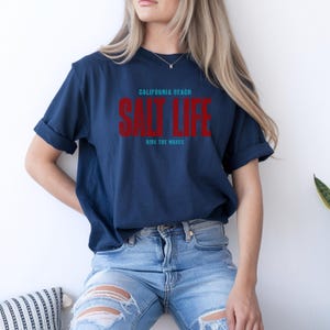 以下が含まれることがあります： 前面に「CALIFORNIA BEACH SALT LIFE RIDE THE WAVES」の文字がプリントされたネイビーブルーのTシャツです。