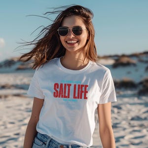 以下が含まれることがあります： 白いTシャツを着た女性が、赤い文字と青い文字で「Salt Life」と書かれています。彼女は砂丘のあるビーチに立っています。