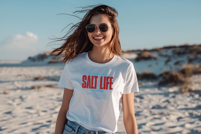 以下が含まれることがあります： 白いTシャツを着た女性が、赤い文字と青い文字で「Salt Life」と書かれています。彼女は砂丘のあるビーチに立っています。