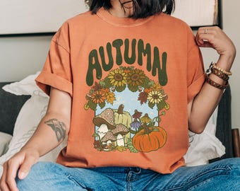 May include: Orange t-shirt with the word "AUTUMN" in green, arched text. The shirt features a graphic of mushrooms, pumpkins, and sunflowers. The shirt is worn with blue jeans.