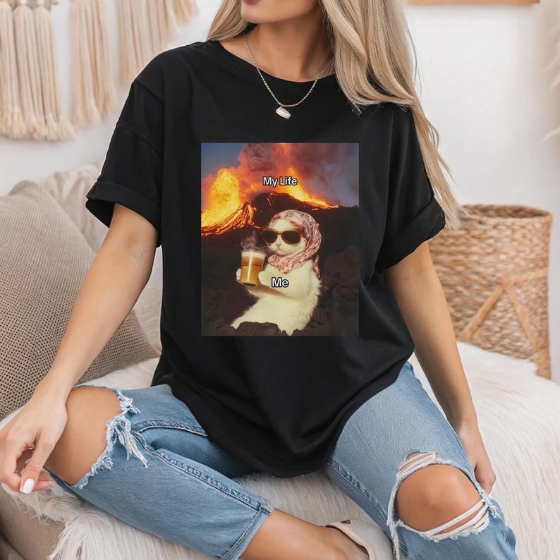 May include: Black t-shirt with a graphic of a cat wearing sunglasses and a headscarf, holding a beer. The text "My Life" is above the cat and "Me" is below the cat. The cat is sitting in front of a volcano.