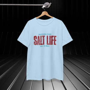 以下が含まれることがあります： ライトブルーのTシャツに、赤と青のグラフィックで「カリフォルニアビーチ ソルトライフ 波に乗ろう」と書かれています。