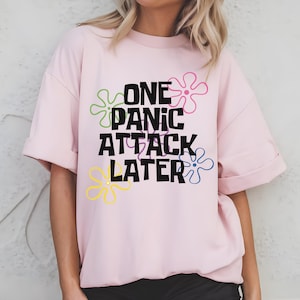 May include: Pink short-sleeved t-shirt with the text "One Panic Attack Later" in black lettering and colourful cartoon flowers.