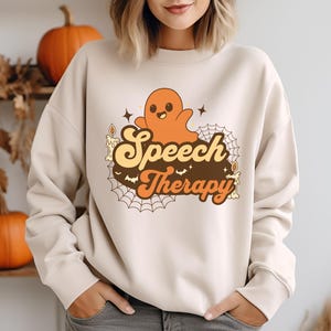 Può includere: Una felpa color crema con una grafica retrò di un fantasma amichevole, candele e ragnatele. Il testo "Speech Therapy" è in lettere arancioni e marroni.