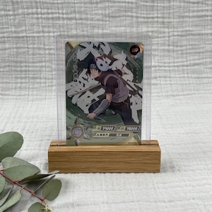 May include: A trading card featuring a character illustration in a protective sleeve, displayed in a wooden stand. The card has a green and white background with Japanese text and character statistics. The stand is made of light brown wood.