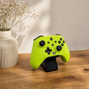 Könnte beinhalten: Ein leuchtend neongelber kabelloser Videospiel-Controller mit schwarzen Analogsticks, einem schwarzen Steuerkreuz und vier farbigen Aktionstasten mit der Beschriftung A, B, X und Y. Der Controller steht auf einer schwarzen Ladestation.
