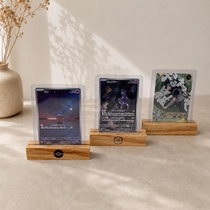 Puede incluir: Tres soportes rectangulares de madera de color marrón claro, cada uno con un pequeño icono circular en blanco y negro en la parte frontal, sostienen tarjetas coleccionables individuales en fundas protectoras transparentes.