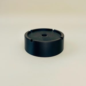 Könnte beinhalten: Ein schwarzer, zylindrischer Aschenbecher mit einem kreisförmigen Design. Der Aschenbecher hat ein zentrales Loch und drei Rillen zum Ablegen von Zigaretten. Der Aschenbecher besteht aus einem mattschwarzen Material.