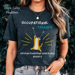 May include: Dark grey heather t-shirt with a graphic of a cat holding crutches. The text on the shirt reads "Occupational Therapy Helping Everyone Reach New Heights".