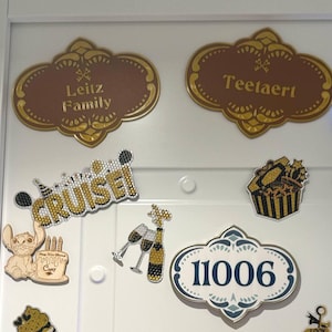 May include: A selection of decorative magnets and plaques on a white surface. Features two brown and gold plaques with family names, a "CRUISE!" magnet, a cake, champagne glasses, a bottle, and a house number plaque.