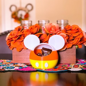 May include: A Mickey Mouse-shaped candy holder with orange, yellow, and white candy corn colors. The holder has white ears and is filled with chocolate candies. The holder is sitting on a table with a colorful table runner and a wooden box filled with orange flowers.