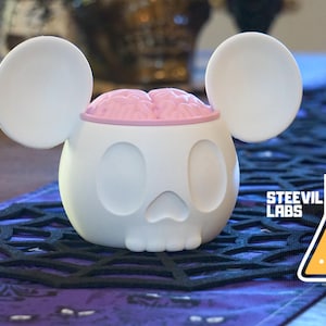 Peut inclure: Crâne en céramique blanche avec un cerveau rose à l'intérieur, en forme d'oreilles de Mickey Mouse. Le crâne est posé sur une surface noire à motif de toile d'araignée.