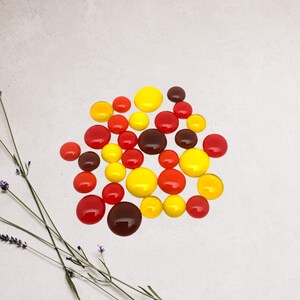 May include: A collection of round, glossy, colorful glass beads in shades of red, yellow, and brown. The beads are arranged in a loose pile on a white surface.