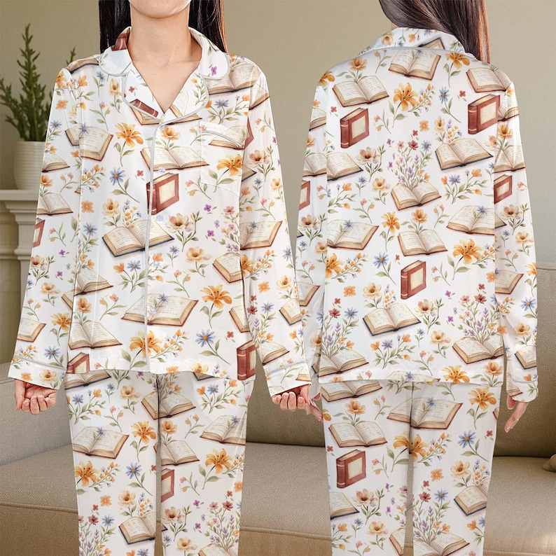 May include: White pyjama set patterned with books and flowers. The long-sleeved top and trousers display open books, orange and yellow flowers, and small blue flowers. The pyjamas have a button-down top with a collar.
