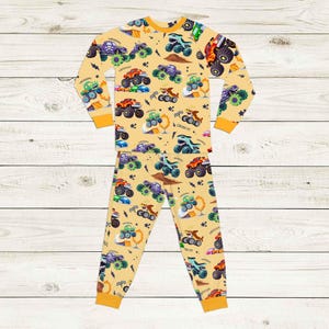 May include: A two-piece pajama set featuring a repeating pattern of colorful monster trucks on a light yellow background. The long-sleeved top and pants have orange trim. The text "VROOOM!" and "CRUSH IT!" are visible.