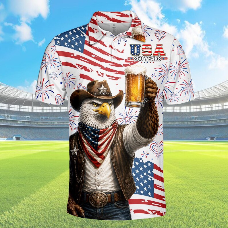 Peut inclure: Polo blanc avec un motif patriotique repr&eacute;sentant un aigle portant un chapeau de cowboy, tenant une bi&egrave;re et un drapeau am&eacute;ricain. Le t-shirt porte le texte "USA 250 YEARS" et un motif de feux d'artifice. Le fond montre un stade et un ciel bleu.