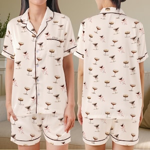 May include: Cream-coloured pyjama set featuring a repeating pattern of martini glasses and pink bows. The short-sleeved top and shorts have dark brown trim. The top has a breast pocket with the name "Olivia" embroidered on it.
