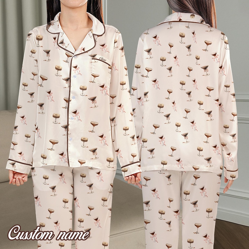 May include: Cream-coloured pyjama set featuring a pattern of martini glasses and pink bows. The long-sleeved top has a notched collar and brown trim. The matching trousers complete the set. The words "Custom name" are written in a script font at the bottom.